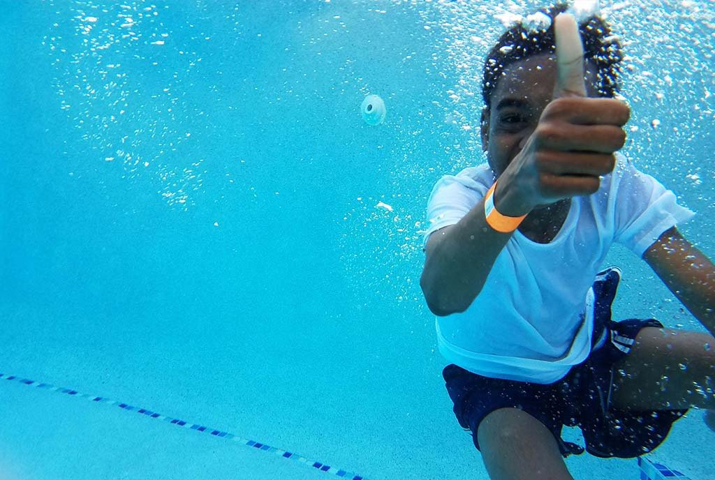 A Black child swims underwater in a pool, holding his thumb up and smiling at the camera.