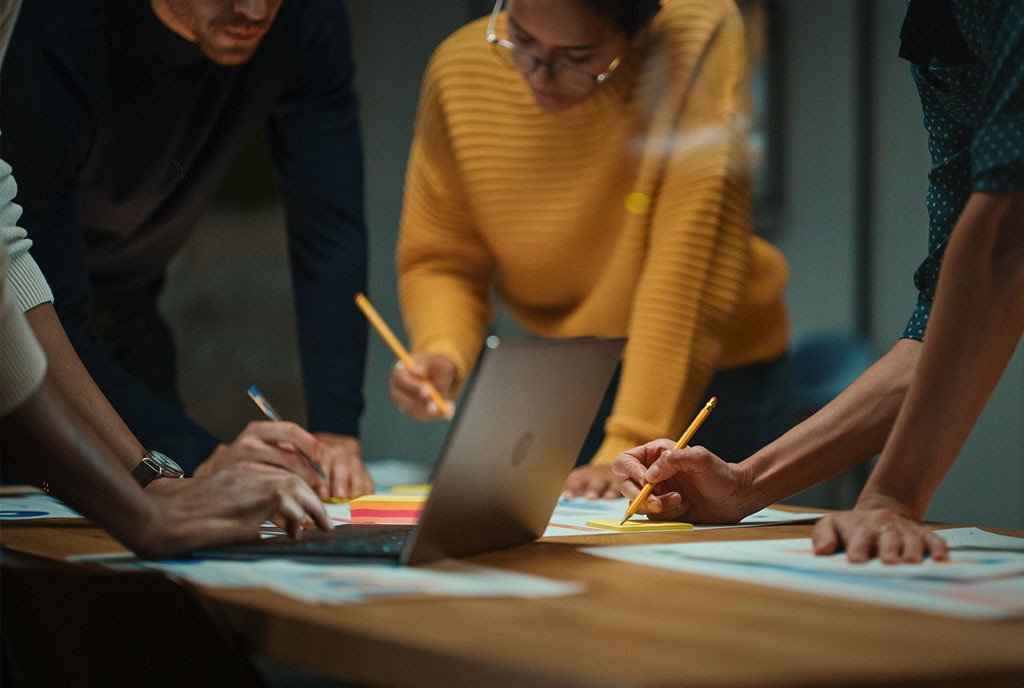 A group of diverse employees in a creative office huddle around a table, planning and writing on post-it notes.