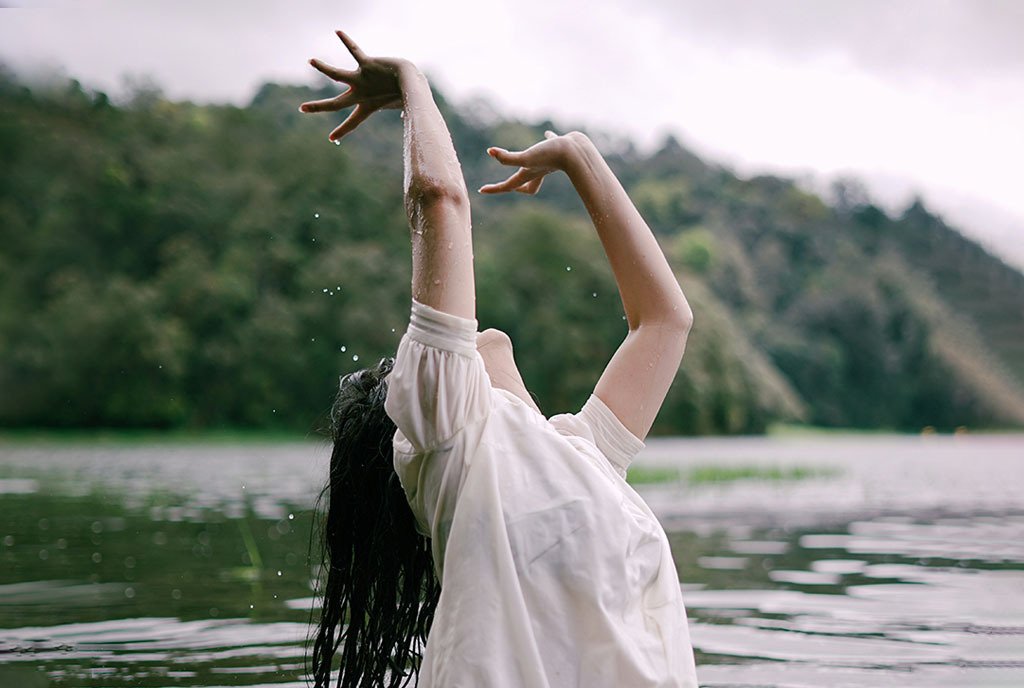 A woman with long black hair emerges from a lake, splashing water as she reaches her arms up. There are trees in the background.