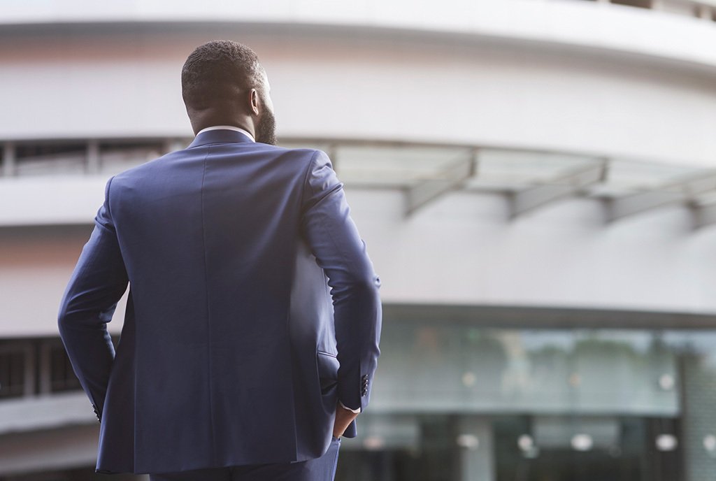A Black man in a suit looks out at an office building with his back towards the camera. His hands are on his hips as if he has a lot to tackle.