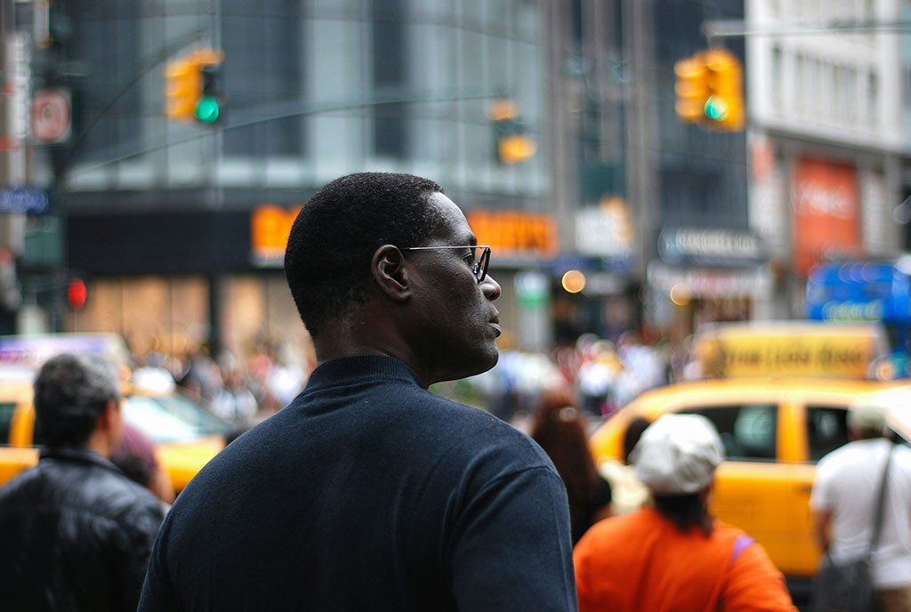 A Black man standing at in intersection in New York city looks over his shoulder with a serious expression on his face.
