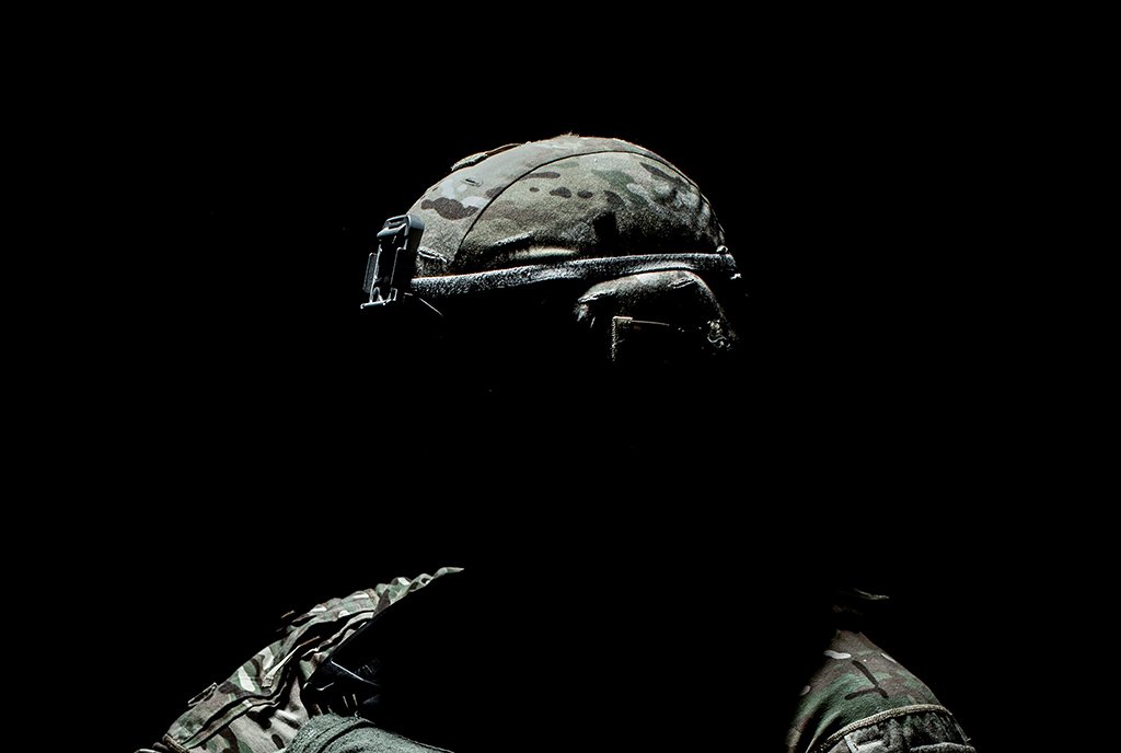 A heavily shadowed person in military fatigues, standing in a dark room.
