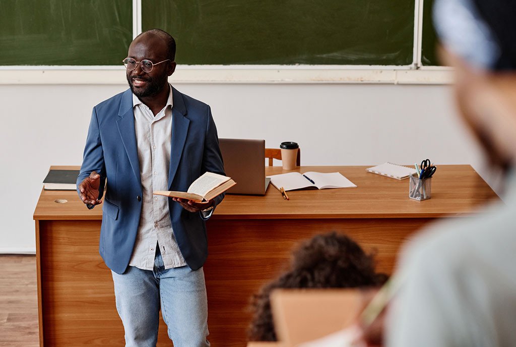 A Black college professor stands at the front of a college classroom in a suit jacket, talking to his students.