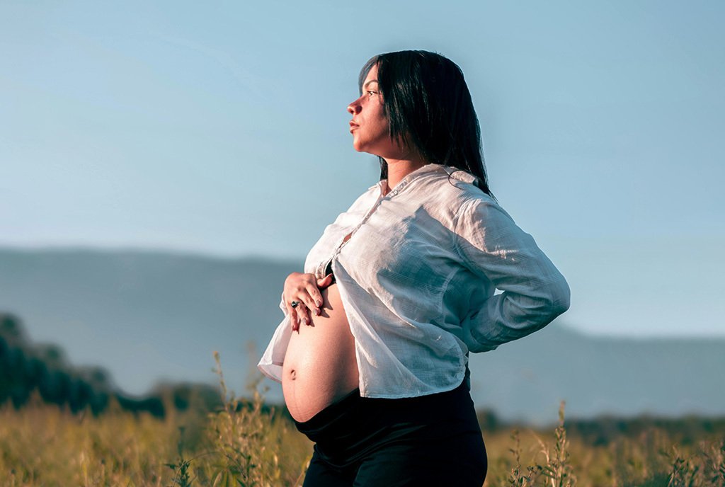 A pregnant women stands in a field holding her exposed belly with a hopeful look on her face.