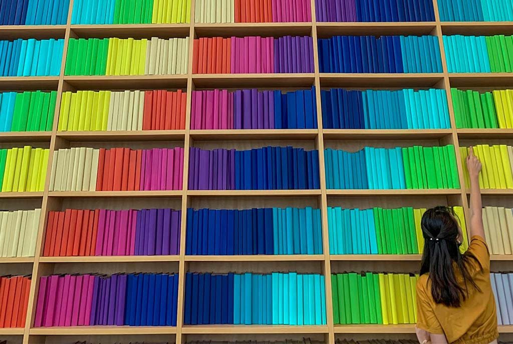 A woman stands in a library of rainbow books, reaching up to grab one.