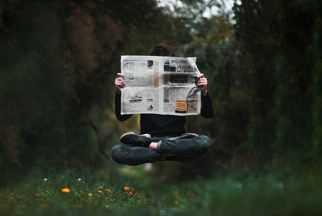 A man holds a newspaper up in front of his face as he floats above a meadow of flowers