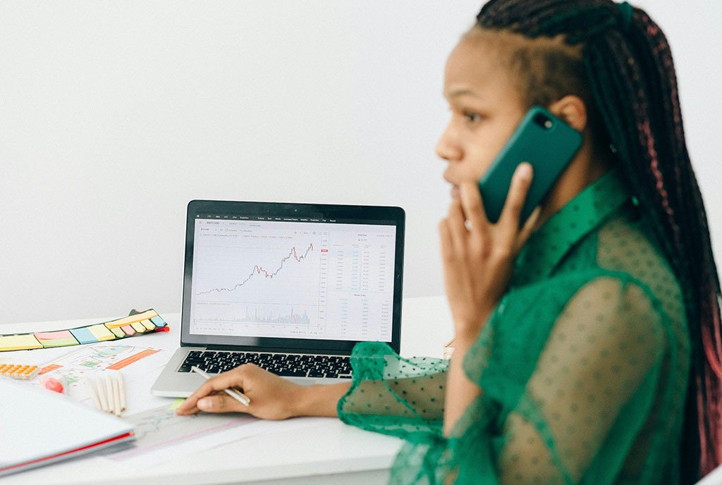 A professional Black woman with a sideshave and braids on the phone. Behind her are a computer and printouts displaying financial charts.