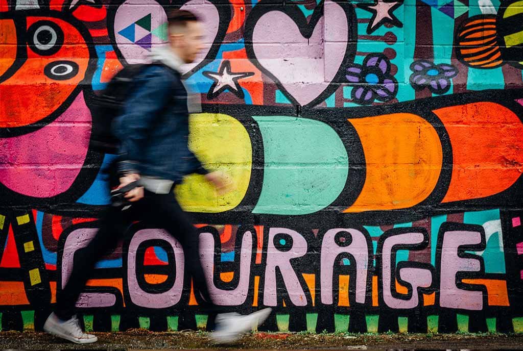 A man walking past a colorful graffiti’d wall that says, “Courage”