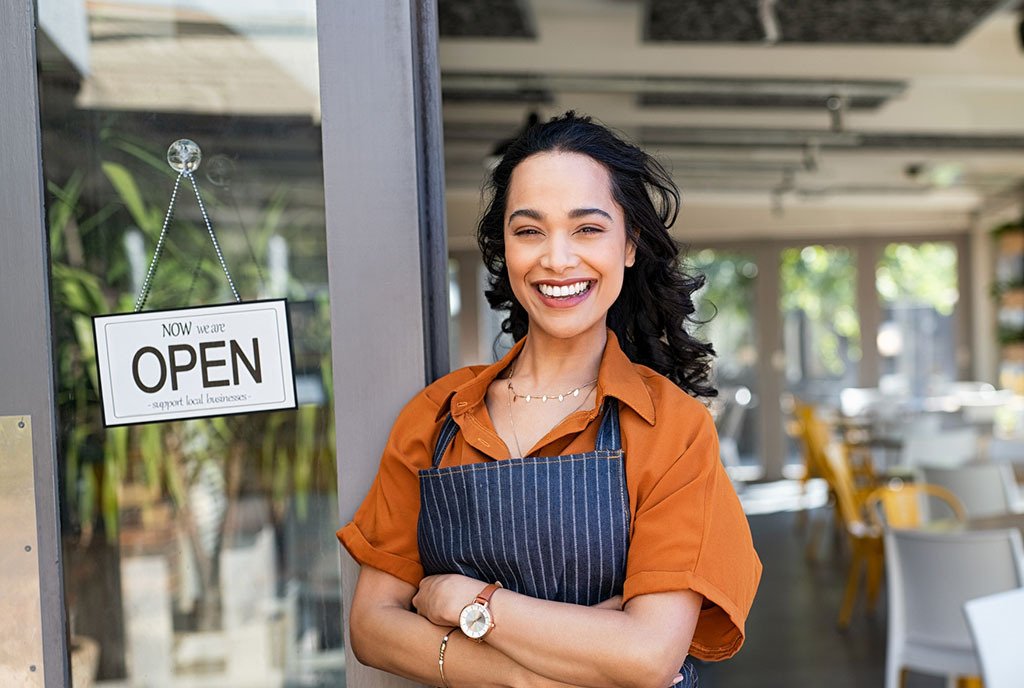 A latinx woman wearing an apron stands at the entrance of her restaurant, next to a sign that reads, “Now we are open. Support local Business”