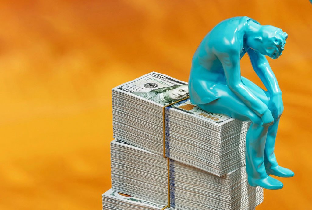 A blue mini figure sits atop a large pile of one hundred dollar bills, hanging his head.