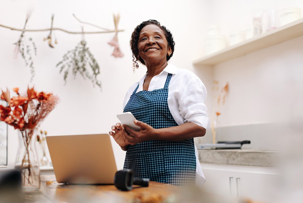 An older Black woman in an apron stands behind the counter of her small business, smiling and looking up optimistically.