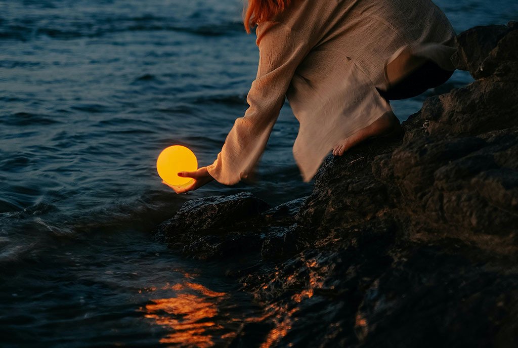 A woman reaches down into a body of water to hold a glowing orb.