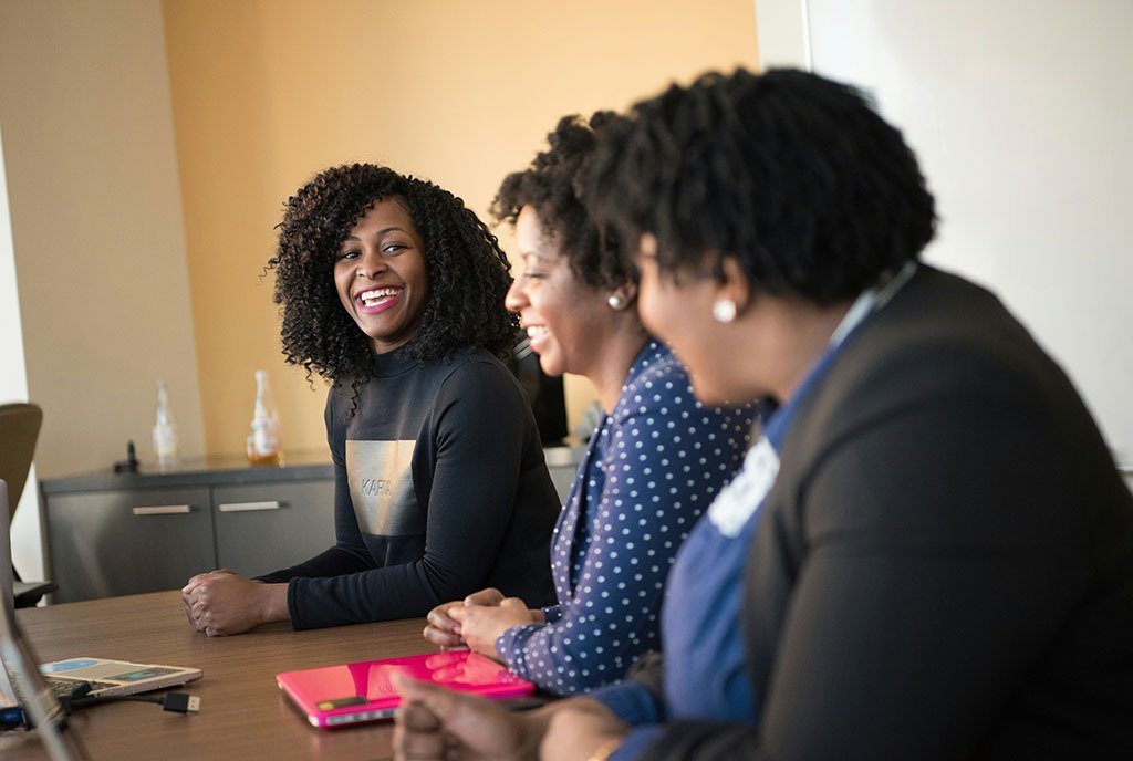 Black women sitting together at a conference table and laughing with each other.