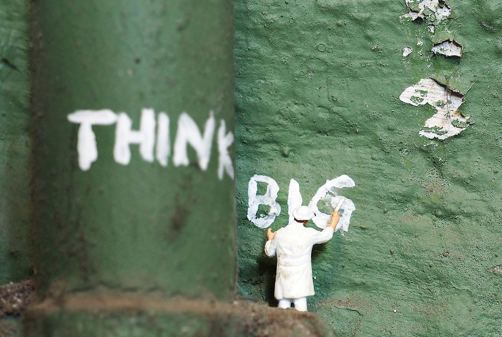 A posed miniture figure painting the words, “Think Big” on a green wall”