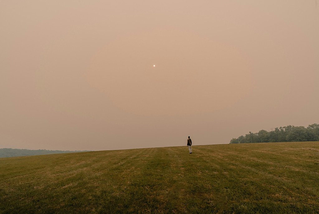 A lone person looks out over a large field at a hazy sky filled with wildfire smoke.