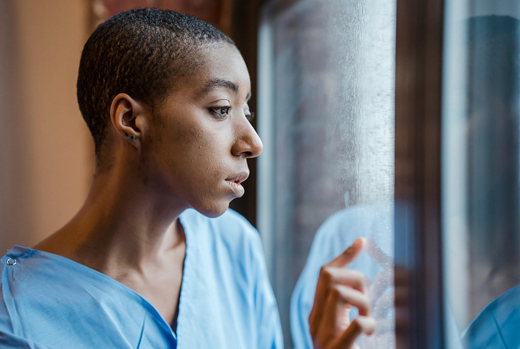 A Black cancer patient with cropped hair peers out of a window, as she wears a blue hospital gown.
