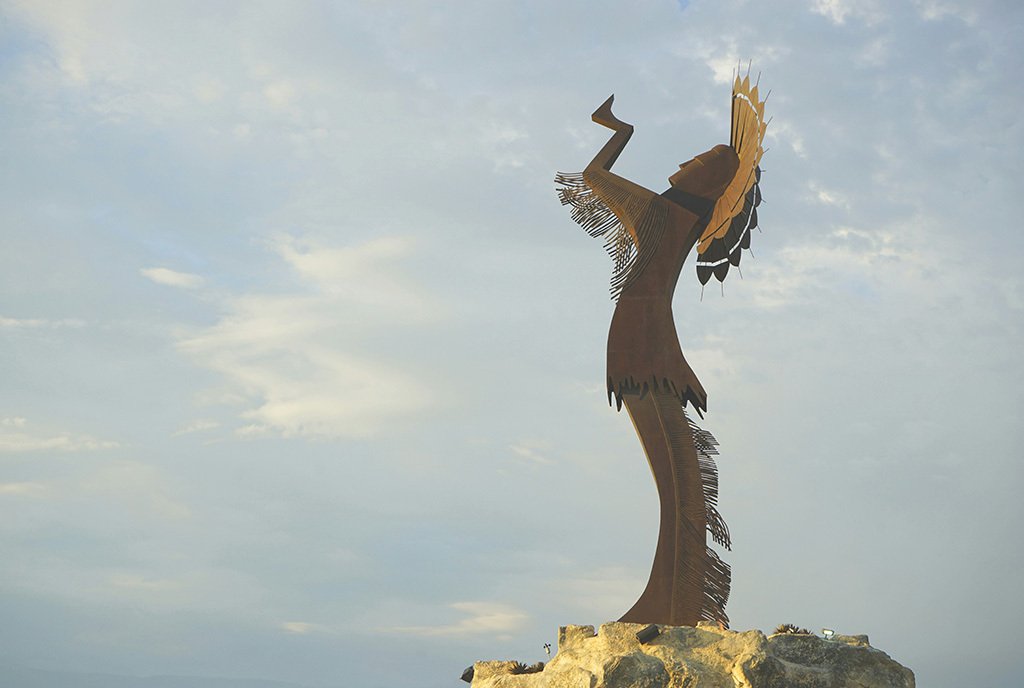 An abstract Sioux Tribe statue of a Native American person wearing a headdress and lifting its hands up to a blue sky.