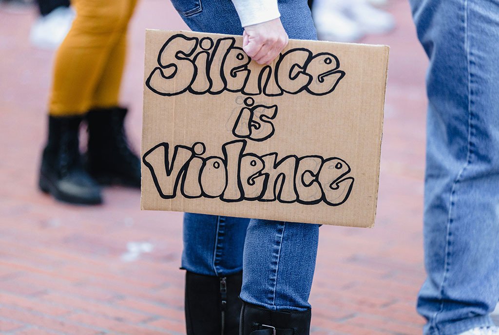 A person holds a cardboard protest sign with bubble letters that read, “Silence is Violence”