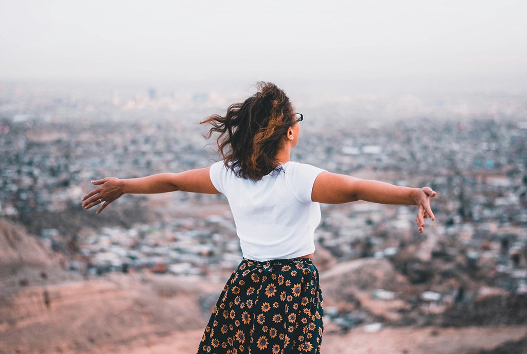 A brown skinned woman with her back to the camera stretches her arms out wide over looking a cliff above a crowded cityscape.