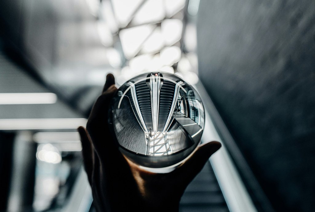 A person holding up a glass orb to a background of down and up escalators. The glass orb inverts the image.