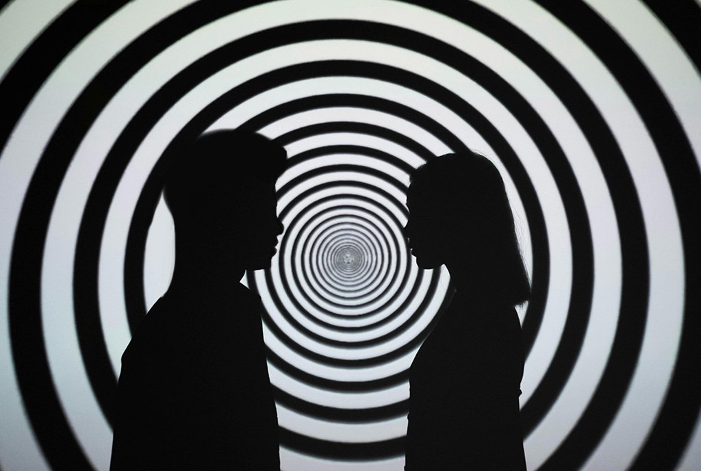 A Black man and White woman facing each other, standing in front of a projected black and white spiral graphic.