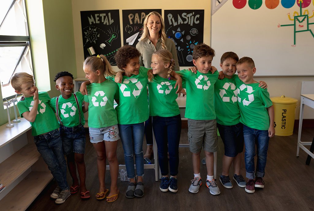 An elementary school teacher stands behind her students, who are learning about recycling and wearing green shirts bearing the recycle symbol.