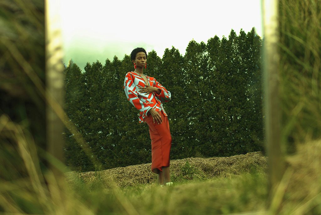 A Black woman in a printed blouse posing in a grassy field, and looking at her own reflection in a proped up mirror.