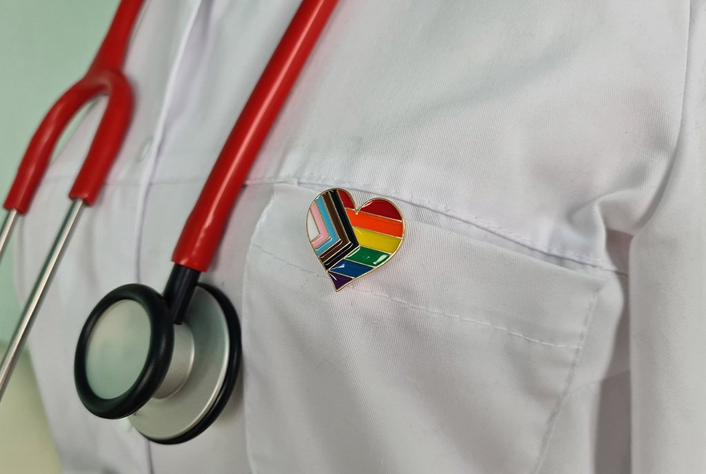 A doctor in a white coat wears a stethoscope and a heart-shaped LGBTQ+ pin on their lapel.