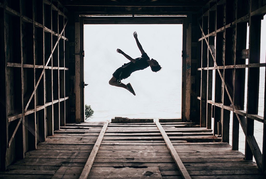 An upward facing view of a person jumping in the opening of a wooden tower. The sky is visible behind him.