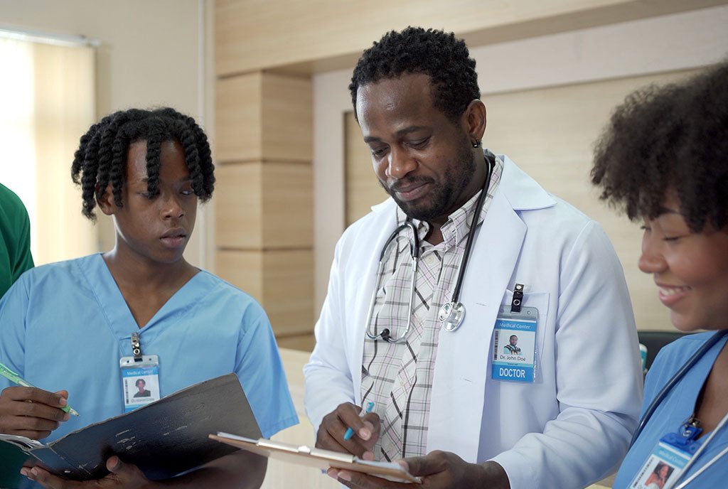 A Black white-coat Doctor stands with two Black medical students, pointing to his clipboard and they listen and take notes.