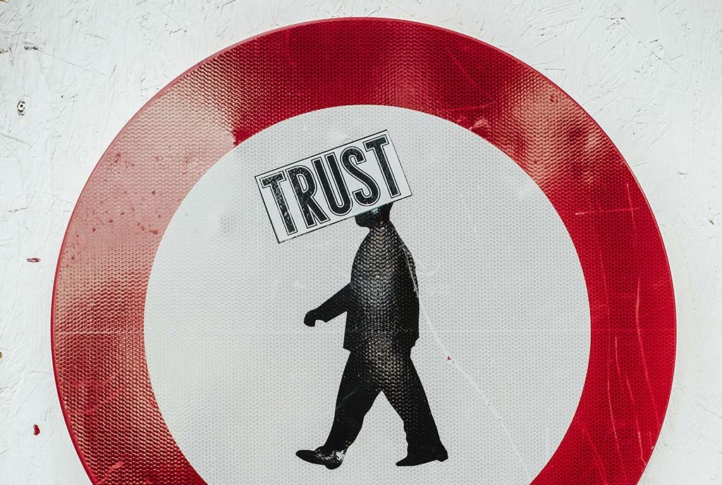 A sign showing a red circle surrounding a man walking. The is a large “Trust” sticker covering the man’s head.