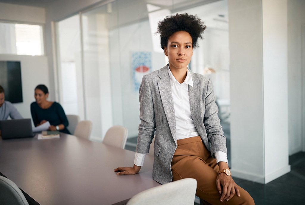 A Black employee sits on a conference room table, as she looks into the camera with a serious expression