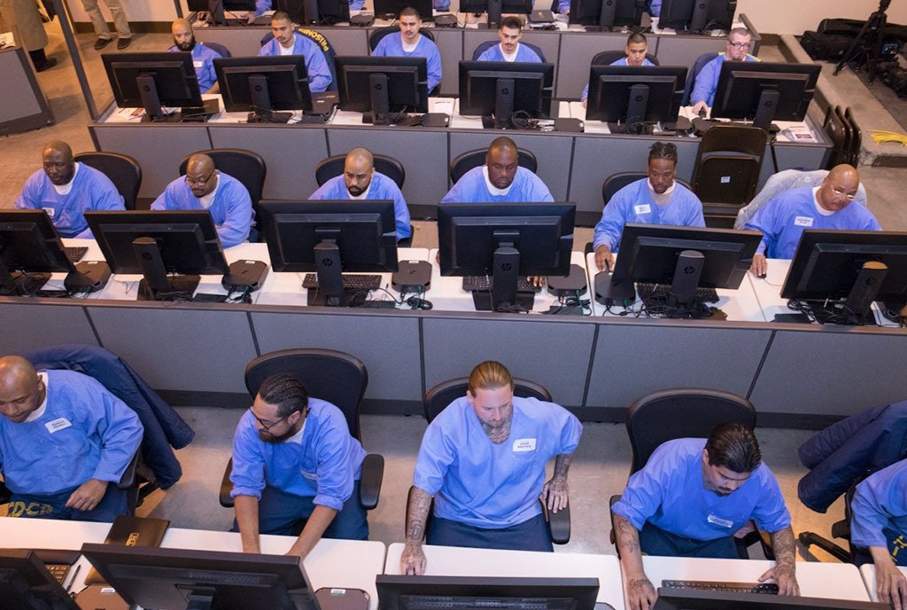A room full of prisoners, or justice-impacted men, in blue jumpsuits using computers to do online learning.