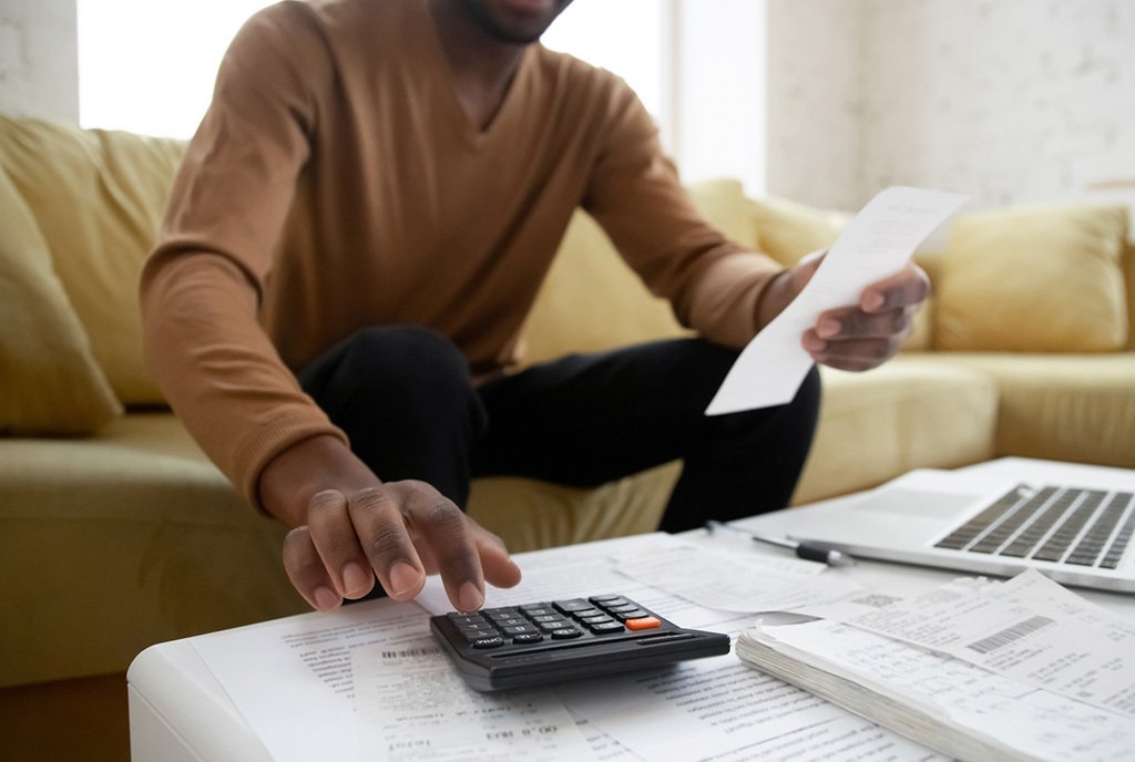 A close-up shot of a Black man doing his taxes, as he plugs numbers into a calculator, with receipts spread out in front of him.