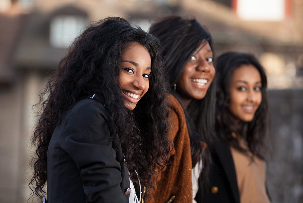 Three teenage girls of color, sitting together in a row and smiling at the camera.