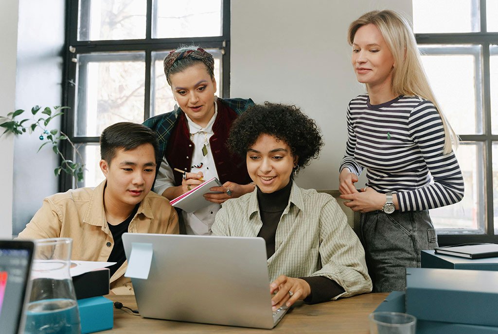 A group of diverse co-workers are gathered around a computer, talking and collaborating as they view the screen together.