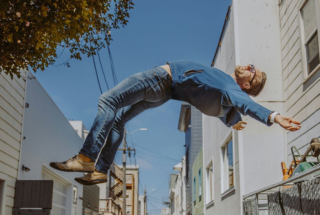 A blond man with glasses jumping up in the air in a bright alleyway and arching his back with the jump.