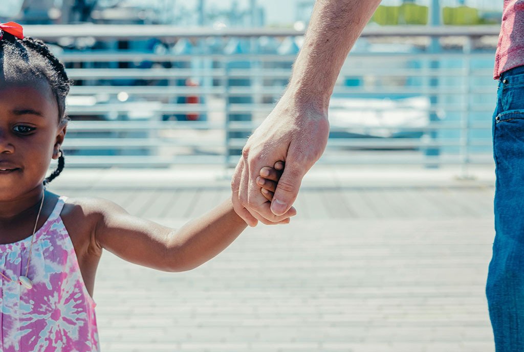 A Black girl toddler wears a pink tie dye dress and ribbon in her hair, as she holds the hand of a white man adult and they walk down a boardwalk.