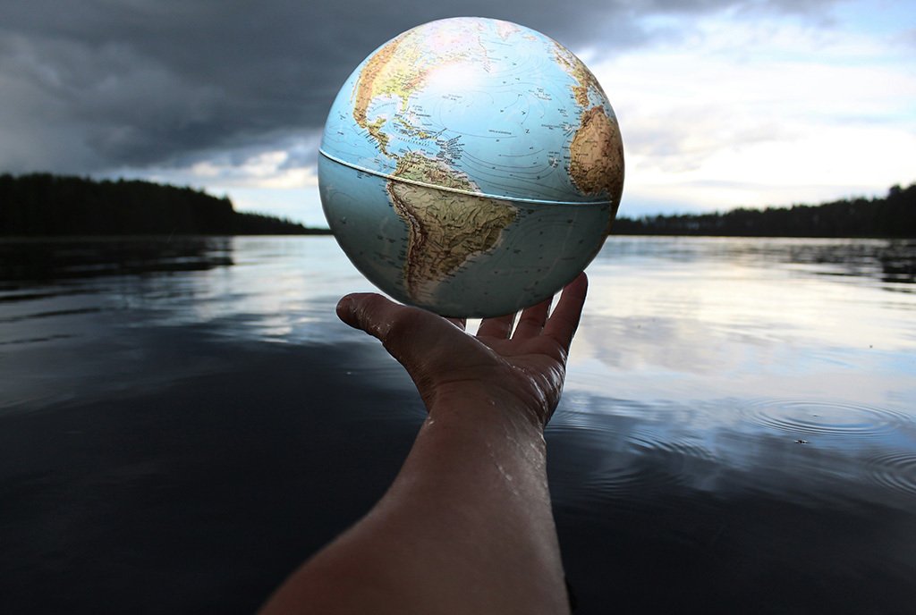 A hands holds a globe of the earth above a body of water surrounded by trees.