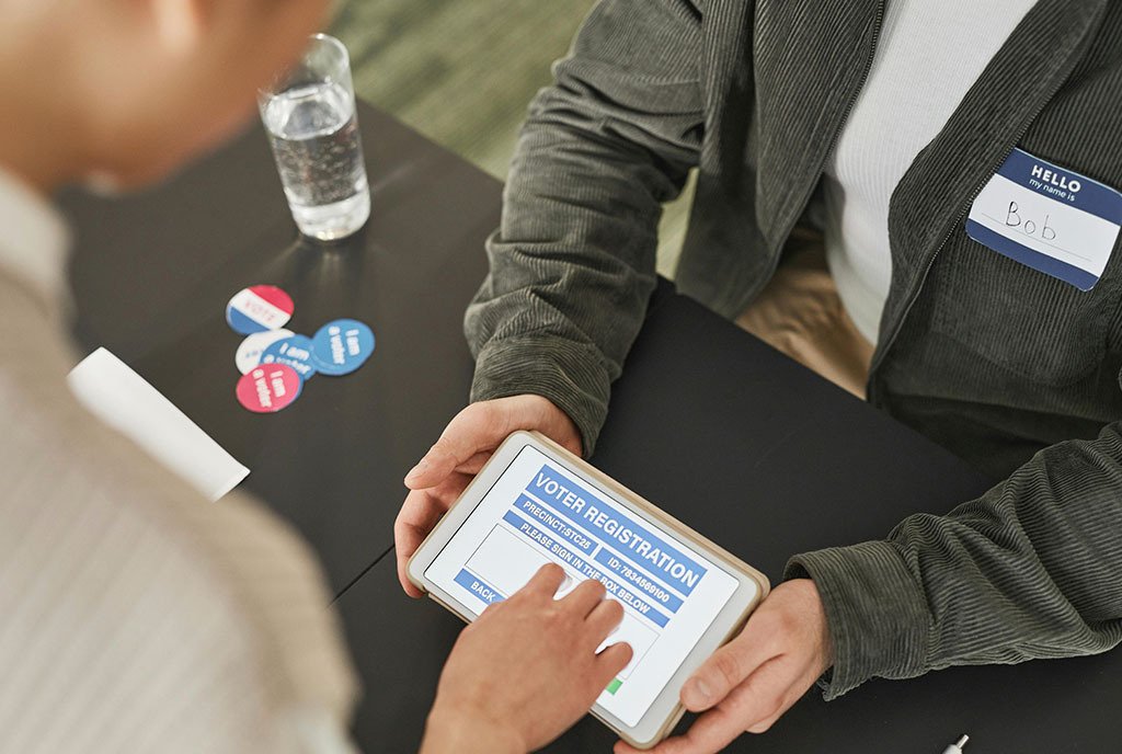 A person sitting at table and presenting a tablet with a voter registration form to a potential voter.