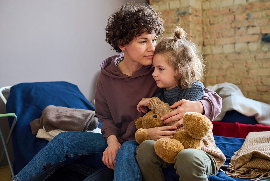 A mother holds her young child close as they sit on the edge of a makeshift bed in a shelter. The child holds a teddy bear.