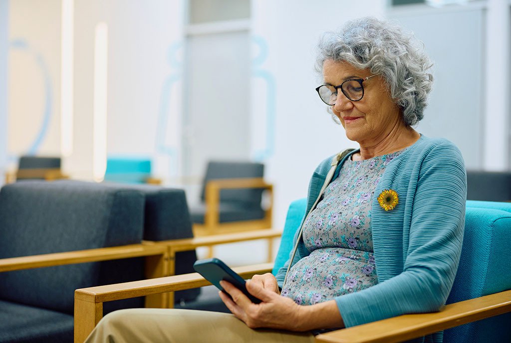 A senior woman sitting in the waiting room of a healthcare provider's room, wearing a hidden disability sunflower pin.