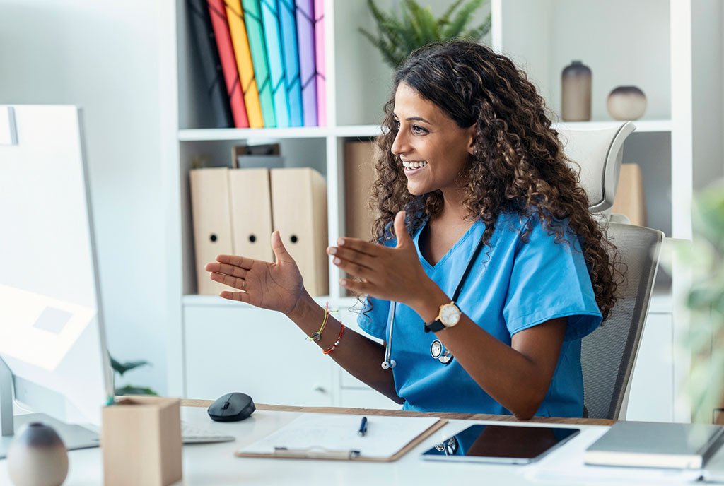 A nurse with long curly hair is seated in blue scrubs in front of a computer screen, where she is gesturing animatedly to someone on a telehealth call.