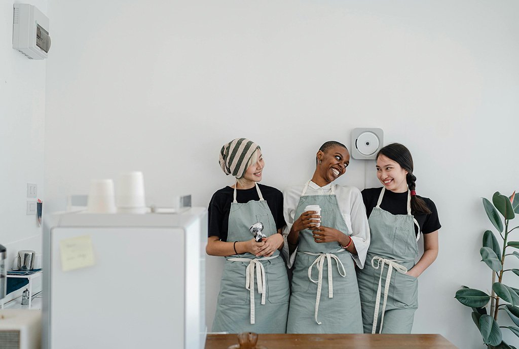 Three diverse woman wearing server aprons, and laughing as the stand against a wall in a small coffee shop.