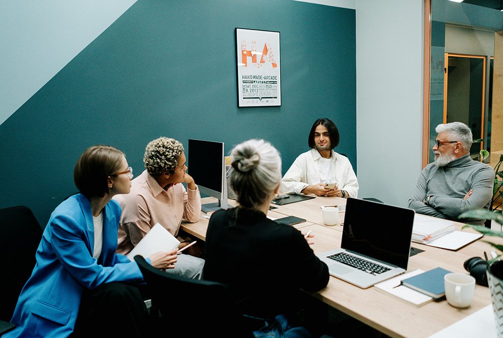 A group of people–two of whom are older and white and three of whom are younger people of color– sitting together. The group looks intently at one of the younger participants, as he begins to speak.