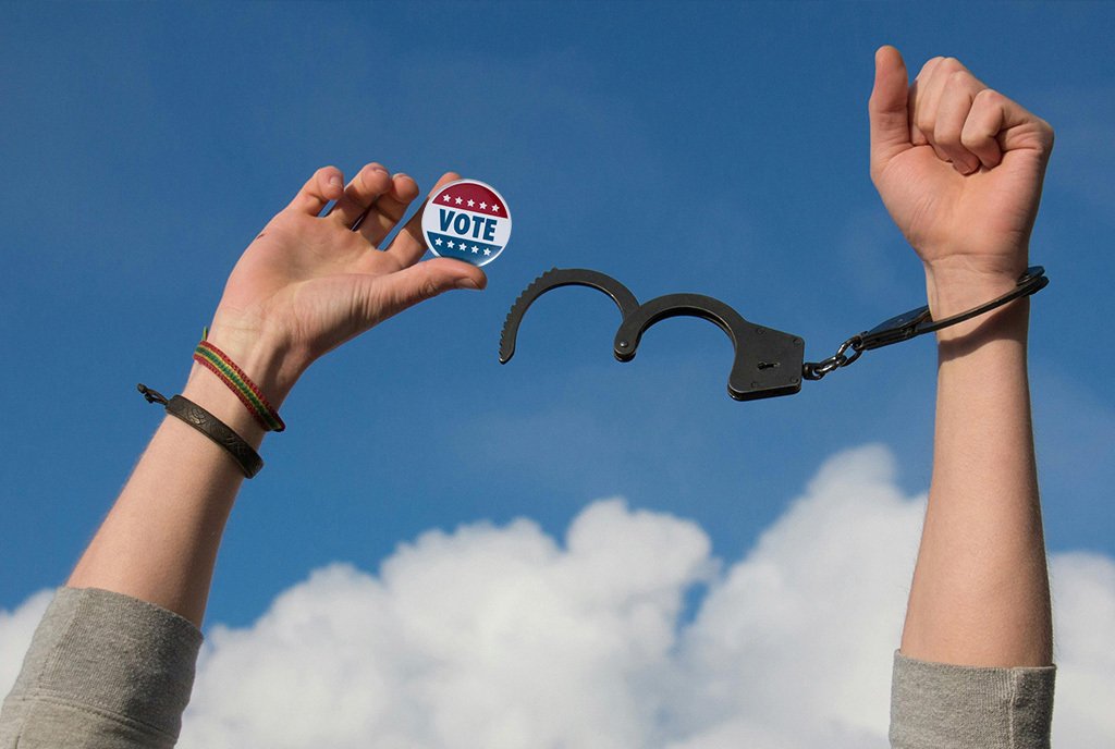 A pair of hands breaking free from handcuffs, and holding a “Vote” pin.