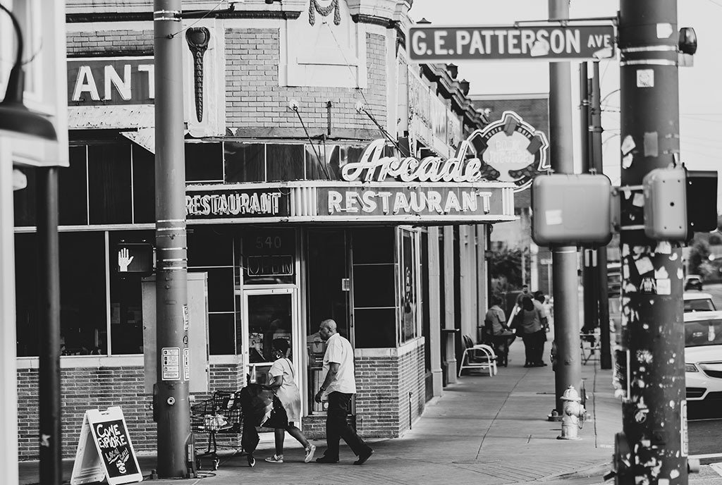 A black and white photo of modern-day downtown Memphis, with Black community members walking up and down the streets. The city’s oldest cafe and a street sign for G.E. Patterson Ave (named after a Black Pentecostal leader) are visible.