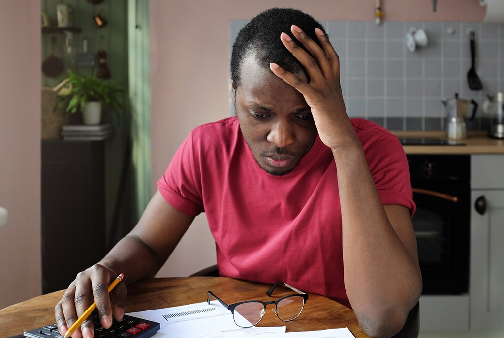 A Black man with a worried look on his face, going through expenses at a desk with a notepad and calculator.