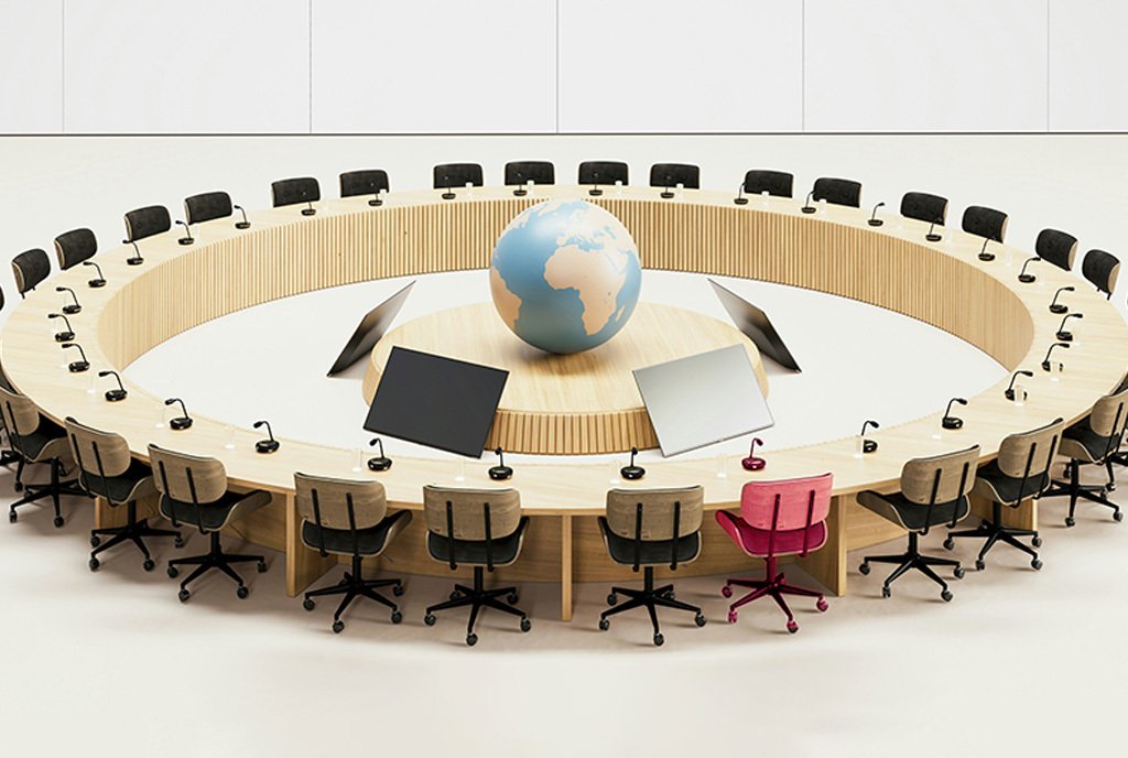 A circular conference table surrounded by empty chairs with microphone in front of them. In the center is a large globe. One of the chairs is bright pink.