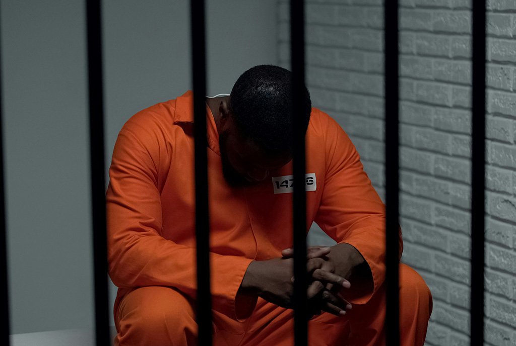 A Black man in an orange jumpsuit hangs his head as he is seated behind bars.